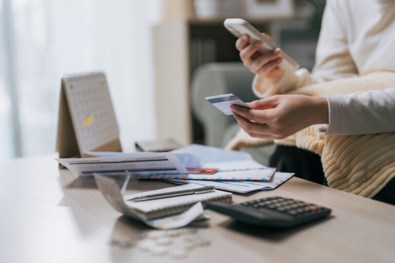 Woman holding credit card and smartphone with unpaid bills on table, representing household debt and extravagant spending lifestyle.