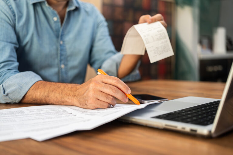Man filling out tax forms at home using a laptop and holding receipts. Financial management and personal accounting concept during tax season